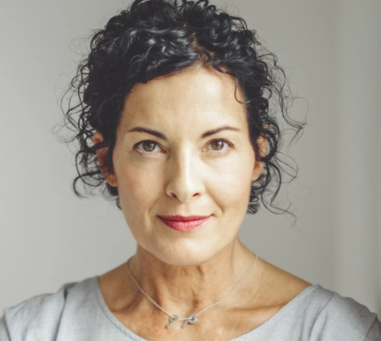 Photo of Eva looking straight into camera, simple background, chain necklace, gray shirt, hair loose at sides of head