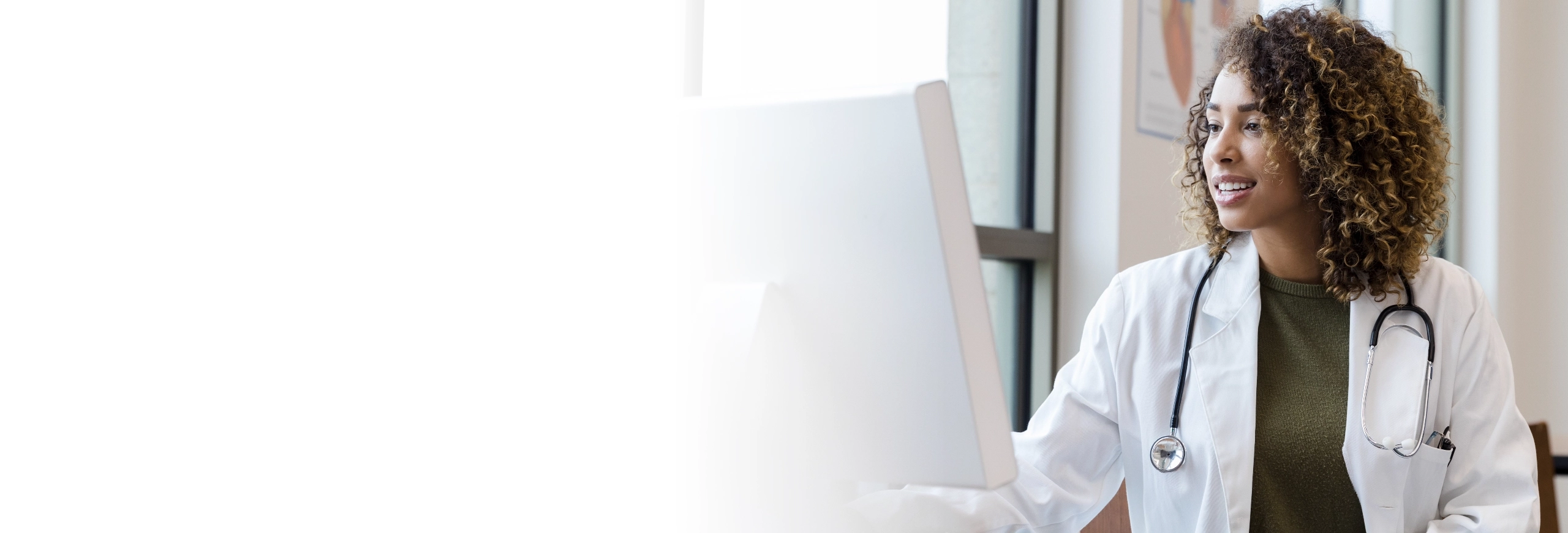 Female healthcare professional wearing white lab coat with stethoscope looking at computer screen.