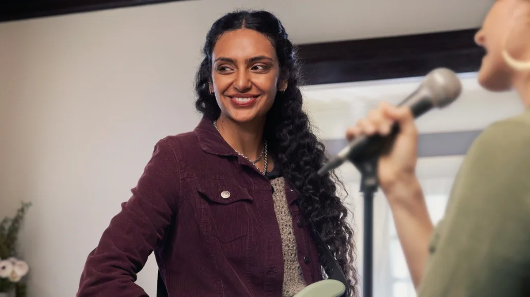 Woman standing and smiling while playing guitar as another woman sings