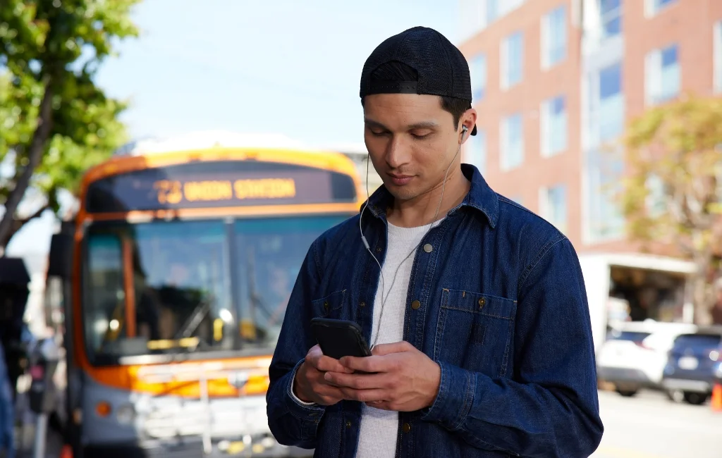 Man wearing earbuds looking down and typing on his phone with a city bus in the background